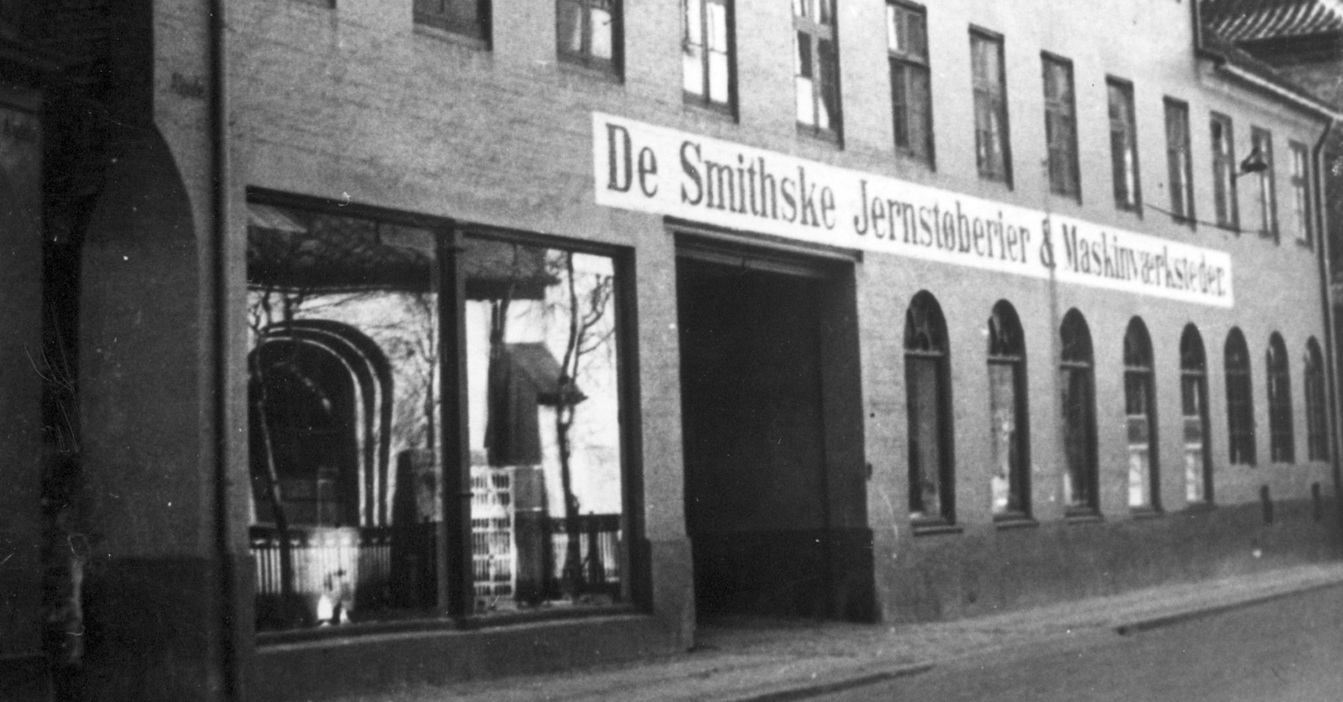 Historic building with "De Smithske Jernstøberier & Maskinværksteder" signage, black and white photo.