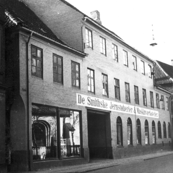 Historic building with "De Smithske Jernstøberier & Maskinværksteder" signage, black and white photo.